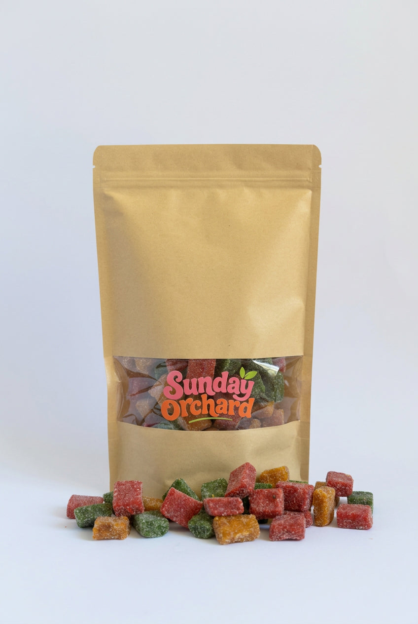 A gold stand-up pouch of Sunday Orchard candy with colourful sugar-dusted fruit cubes spilled in front on a white background.