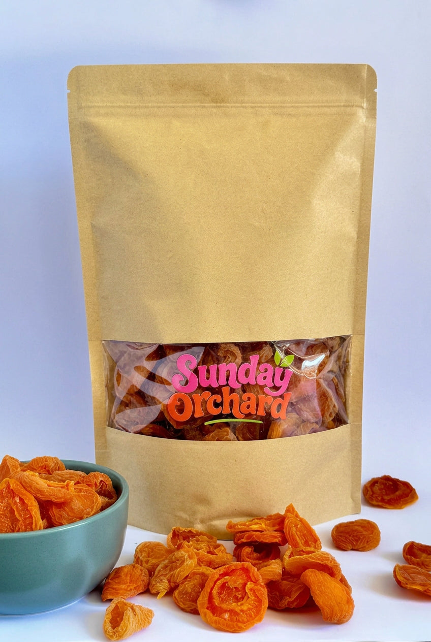 A kraft paper stand-up pouch labeled 'Sunday Orchard' filled with dried apricots, shown on a white background with a teal bowl and scattered apricots in front.