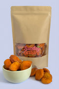 A kraft paper stand-up pouch with a clear window showing dried apricots inside, with a small white bowl and several loose dried peaches in front on a white background. The pouch has a visible 'Sunday Orchard' logo on the window. 