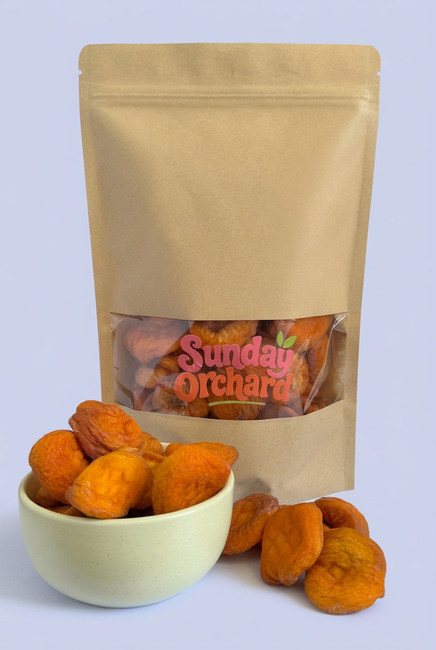 A kraft paper stand-up pouch with a clear window showing dried apricots inside, with a small white bowl and several loose dried peaches in front on a white background. The pouch has a visible 'Sunday Orchard' logo on the window. 