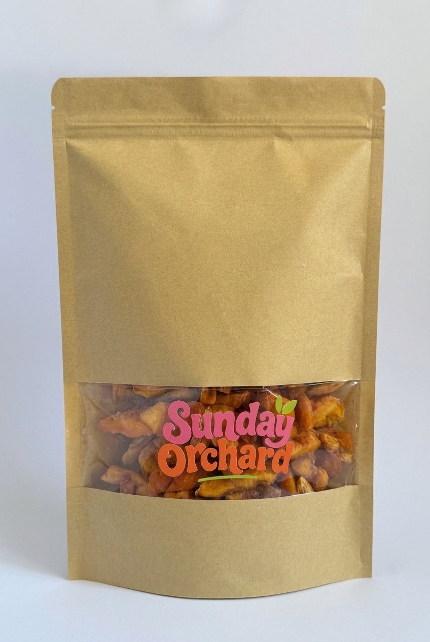 A gold stand-up pouch of snack mix with a clear window showing orange and yellow pieces inside. The front has a pink and orange 'Sunday Orchard' logo, photographed on a plain white background.