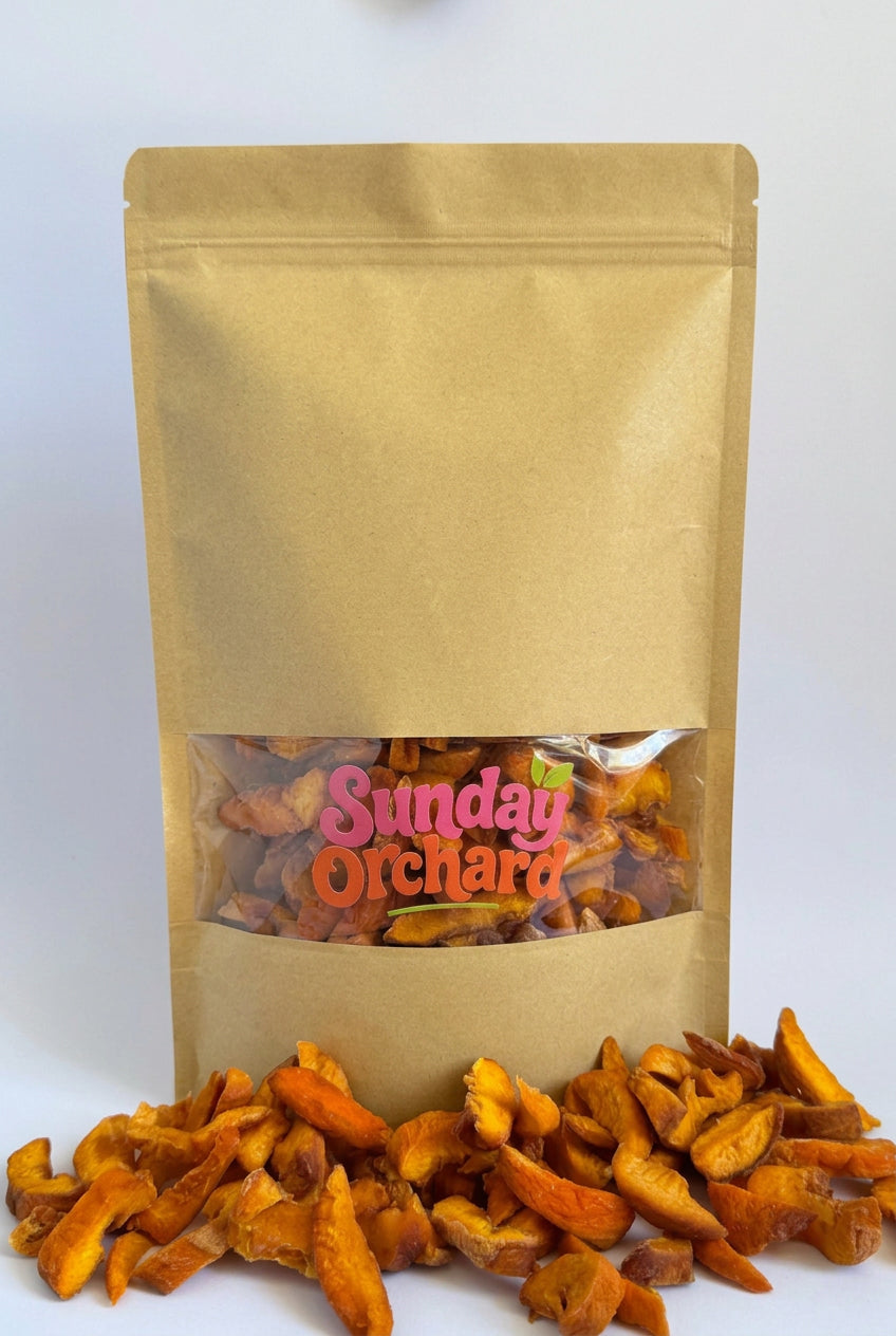 A kraft paper stand-up pouch with a clear window showing dried peach fruit slices inside, with more dried peach fruit scattered in front on a white background. The pouch has a visible 'Sunday Orchard' logo on the window.