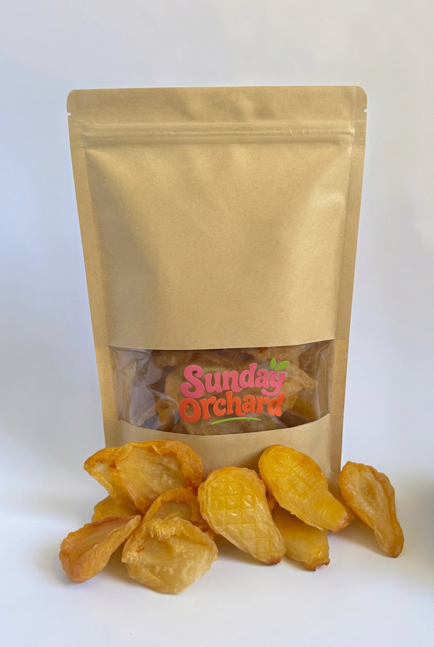A stand-up kraft paper pouch with a clear window labeled 'Sunday Orchard' containing dried pears, with several golden dried pear pieces placed in front on a white background.