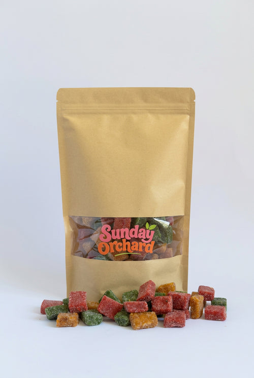 A gold stand-up pouch of Sunday Orchard candy with colourful sugar-dusted fruit cubes spilled in front on a white background.