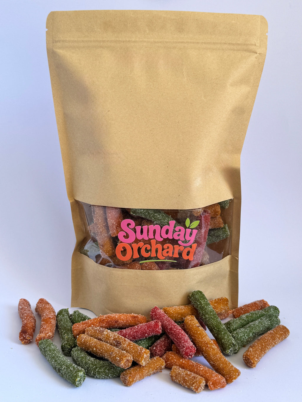 A kraft paper stand-up pouch with a clear window showing colorful fruit sticks inside, with several candy pieces spilled in front on a white background. The pouch label reads 'Sunday Orchard'.