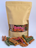 A kraft paper stand-up pouch with a clear window showing colorful fruit sticks inside, with several candy pieces spilled in front on a white background. The pouch label reads 'Sunday Orchard'.