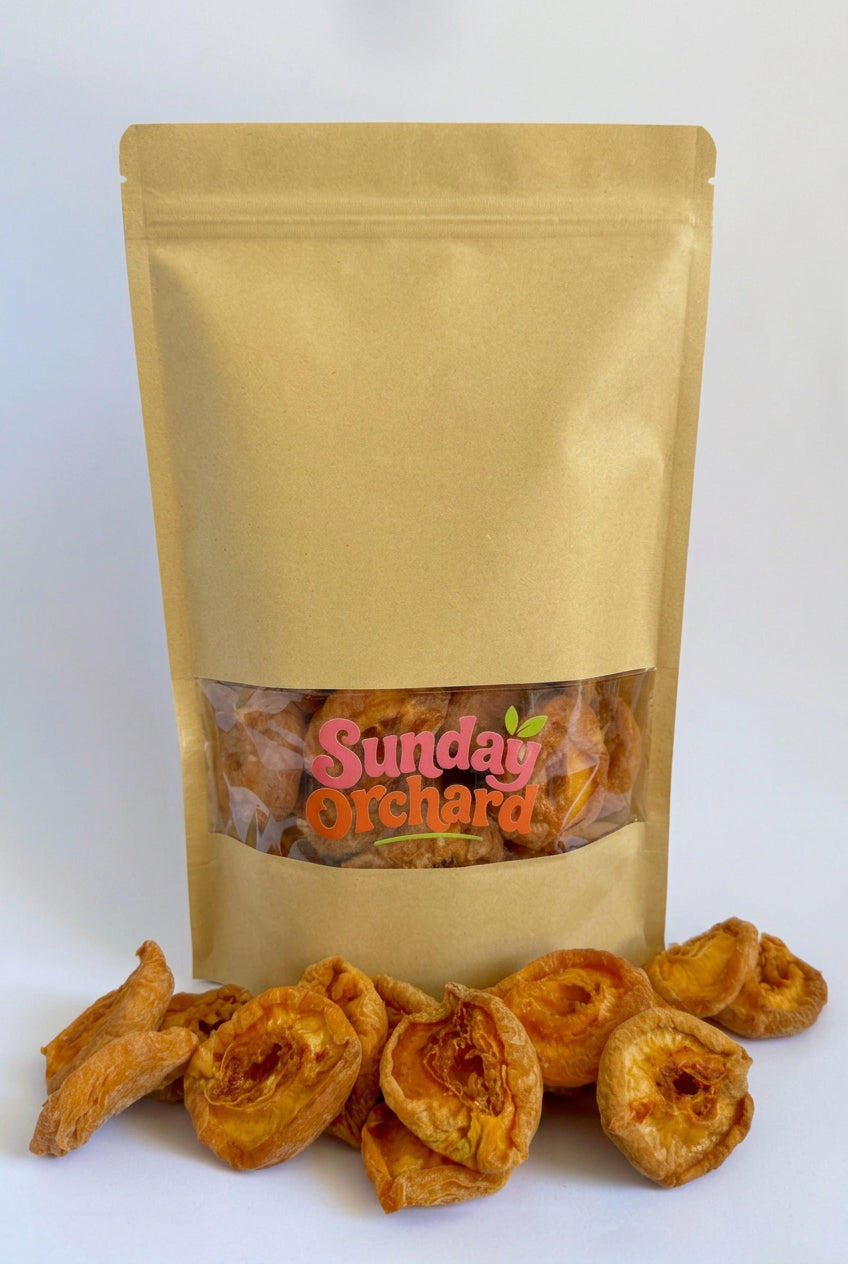 A kraft paper stand-up pouch with a clear window showing dried peaches inside, with several dried peach pieces spilled in front on a white background. The pouch has a visible 'Sunday Orchard' logo.