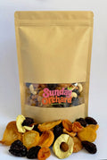A kraft paper stand-up pouch labeled 'Sunday Orchard' filled with mixed dried fruit, with loose dried apple slices, apricots, pears, peaches and prunes arranged in front on a white background.