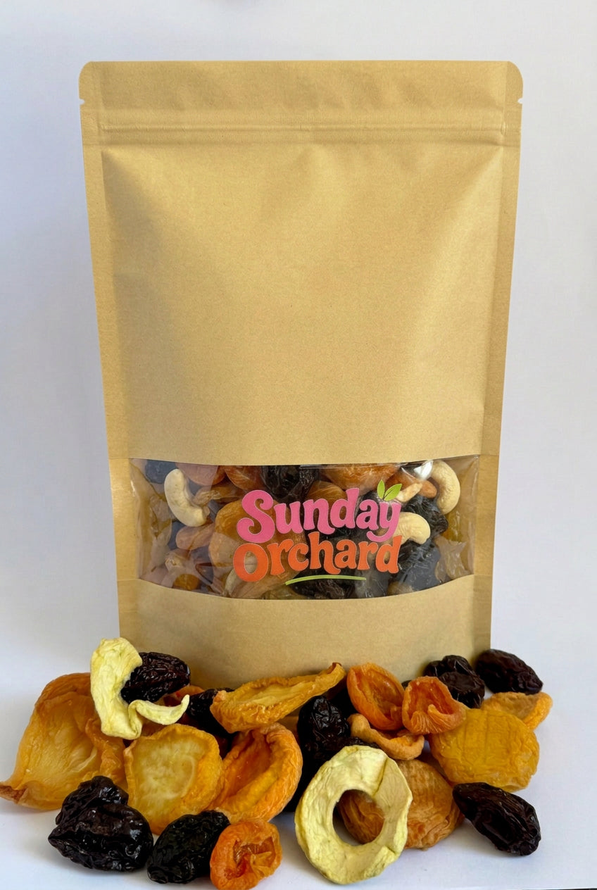 A kraft paper stand-up pouch labeled 'Sunday Orchard' filled with mixed dried fruit, with loose dried apple slices, apricots, pears, peaches and prunes arranged in front on a white background.