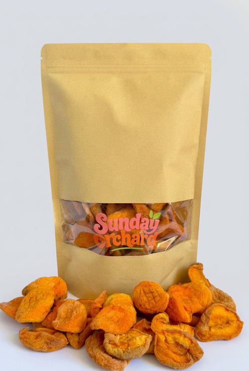 A kraft paper stand-up pouch with a clear window showing dried apricots inside, with additional dried peaches spilled in front on a white background. The pouch has a visible 'Sunday Orchard' logo on the clear window.